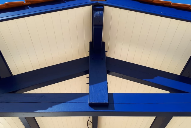 Charpente en bois apparente peinte en bleu soutenant un avant-toit sous un ciel bleu.