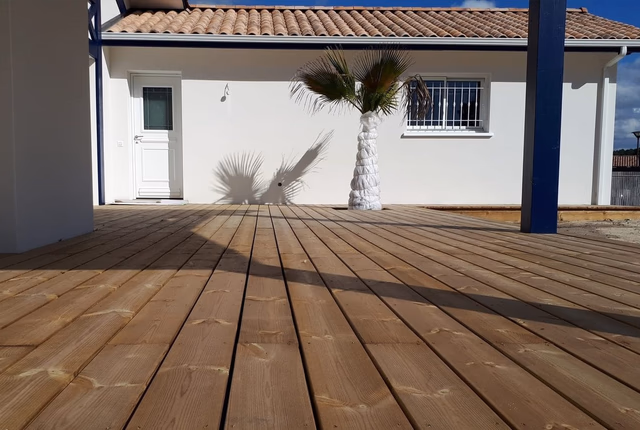 Grande terrasse en bois clair devant la façade blanche d'une maison avec un palmier en pot.