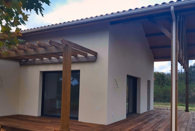 Façade claire avec pergola en bois, grandes baies vitrées et terrasse en lames de bois à Saint-Paul-en-Born.