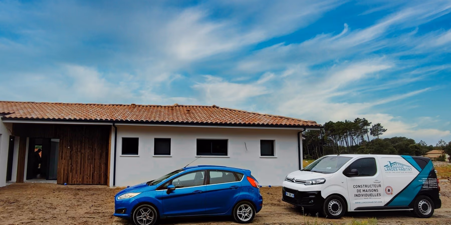 Voiture bleue et utilitaire blanc de constructeur stationnés devant une maison neuve aux tuiles rouges.