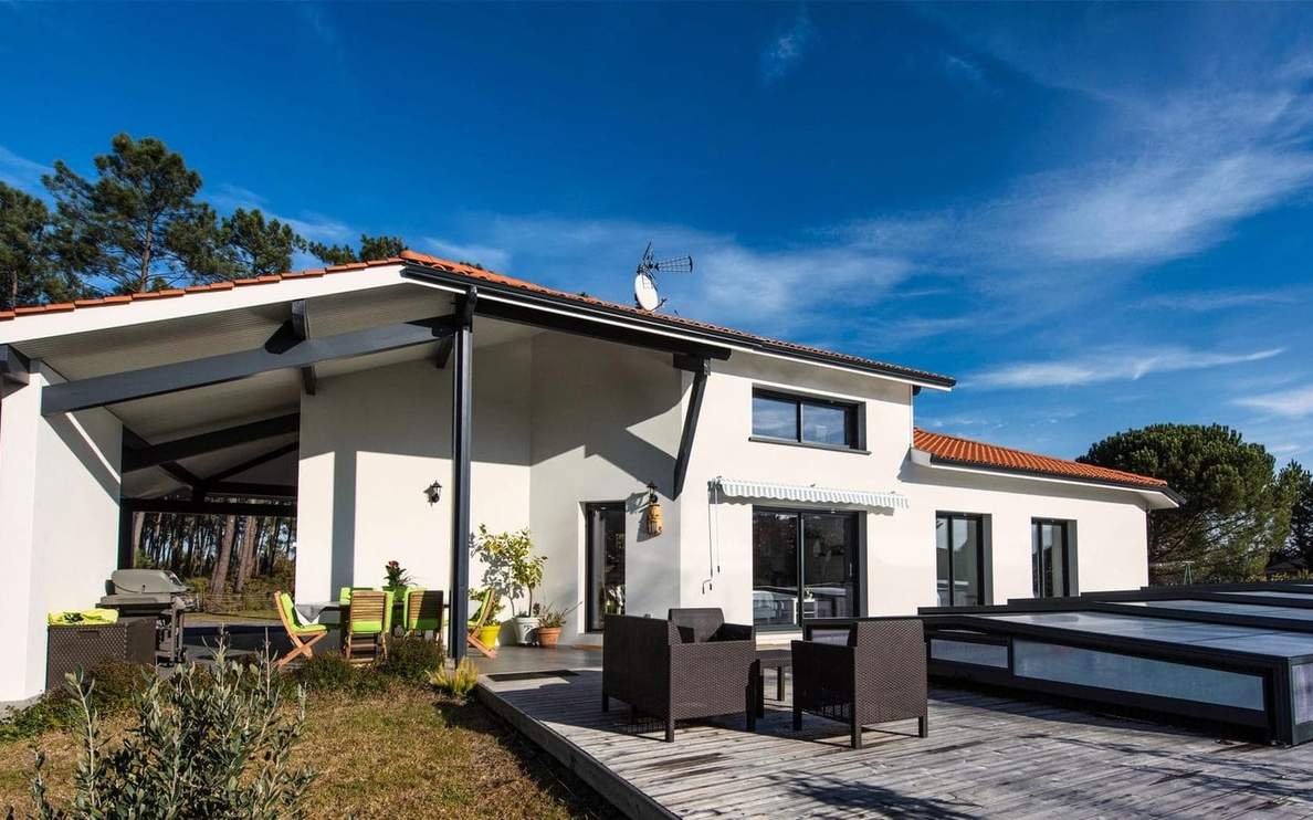 Façade blanche d'une maison contemporaine avec terrasse en bois, mobilier de jardin et abri couvert.