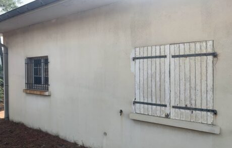 Mur extérieur d'une maison avec une fenêtre grillagée et des volets en bois fermés.